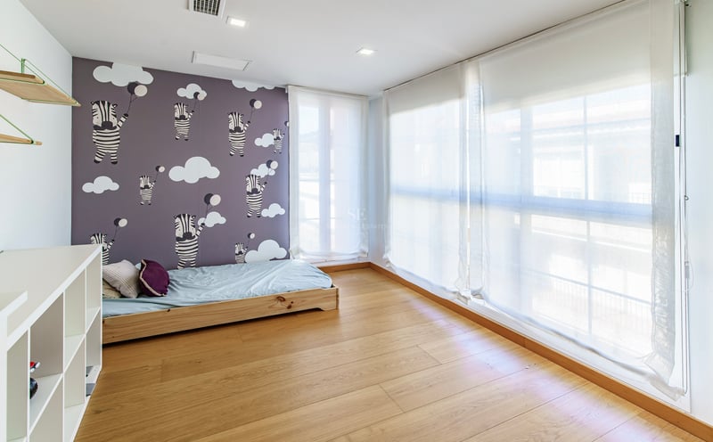 Ljust barnrum med zebramönstrad tapet, ljust trägolv och stora fönster med vita tunna gardiner.
