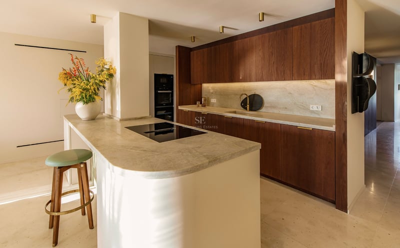 Moderne Küche mit dunklen Holzschränken, Naturstein-Arbeitsplatten und einer geschwungenen Insel mit Barhocker.