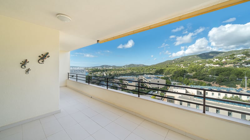 Spacious white tiled balcony with a black metal railing overlooking green hills and a blue sky with white clouds.