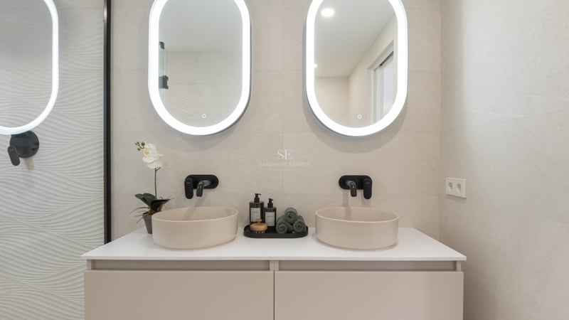Salle de bain moderne avec deux vasques à poser, robinetterie noire et miroirs ovales rétroéclairés sur mur beige.