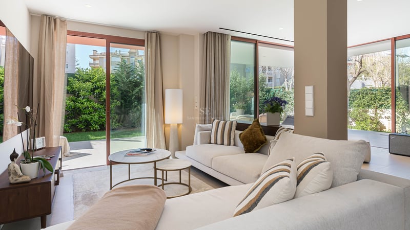 Bright modern living room with cream sofa, marble tables, and floor-to-ceiling glass doors opening to a lush garden.