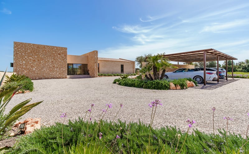 Moderne Villa mit Natursteinwänden, großer Kiesauffahrt und Carport unter strahlend blauem Himmel.