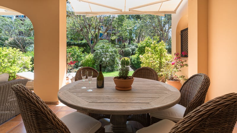 A covered terrace with a round wooden table, wicker chairs, and a view of a lush Mediterranean garden.