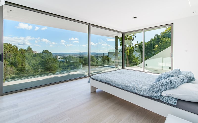 Chambre moderne avec lit blanc et grandes baies vitrées donnant sur une forêt verdoyante et la ville au loin.
