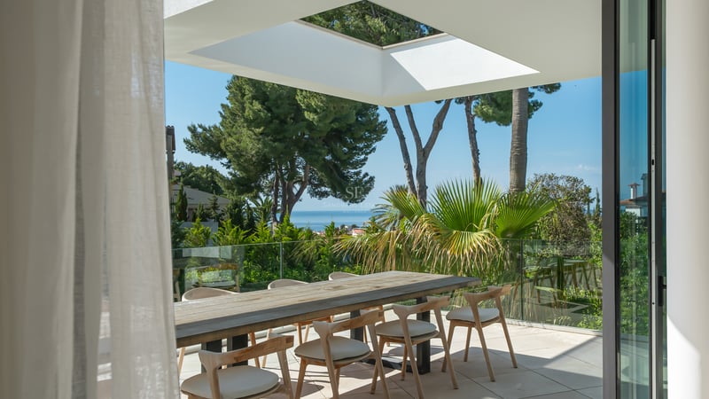 Espace repas extérieur moderne sur une terrasse avec table en bois, garde-corps en verre et vue sur la mer.