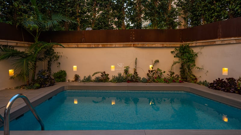 Piscine rectangulaire moderne au crépuscule avec eau turquoise, appliques murales chaleureuses et végétation tropicale.