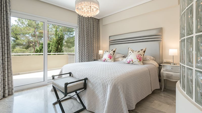 Bright master bedroom with marble floors, silver headboard, and sliding glass doors opening to a terrace with tree views.