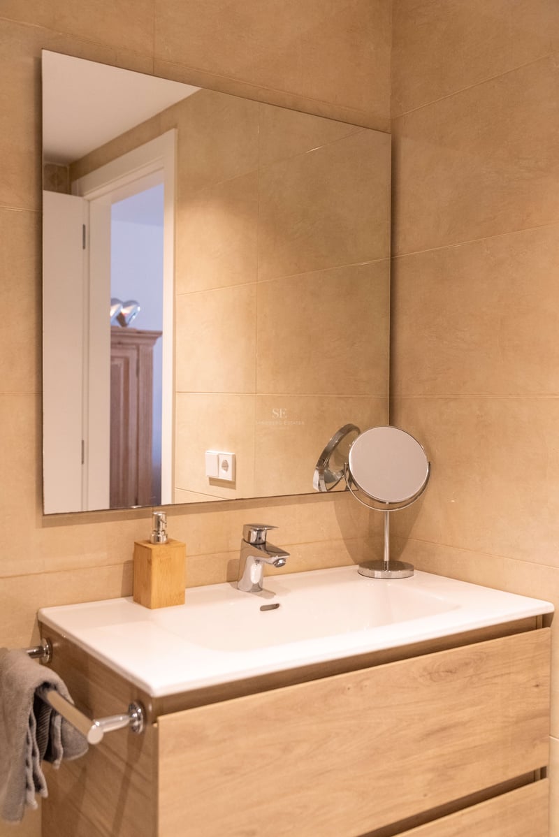 Weißes Keramikwaschbecken auf hellem Holzunterschrank, großer Spiegel und sandfarbene Wandfliesen.