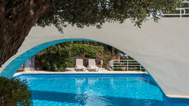 Blue swimming pool and sun loungers framed by a white architectural arch and lush Mediterranean greenery.