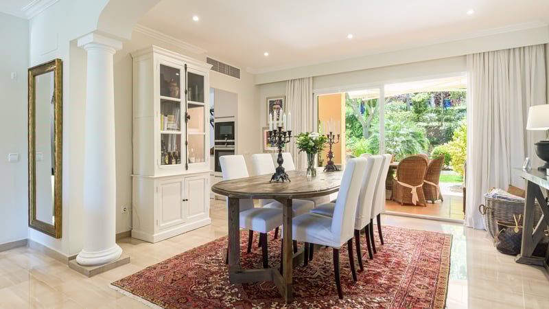 Dining room featuring an oval wooden table, white upholstered chairs, and an oriental rug, overlooking a lush garden.