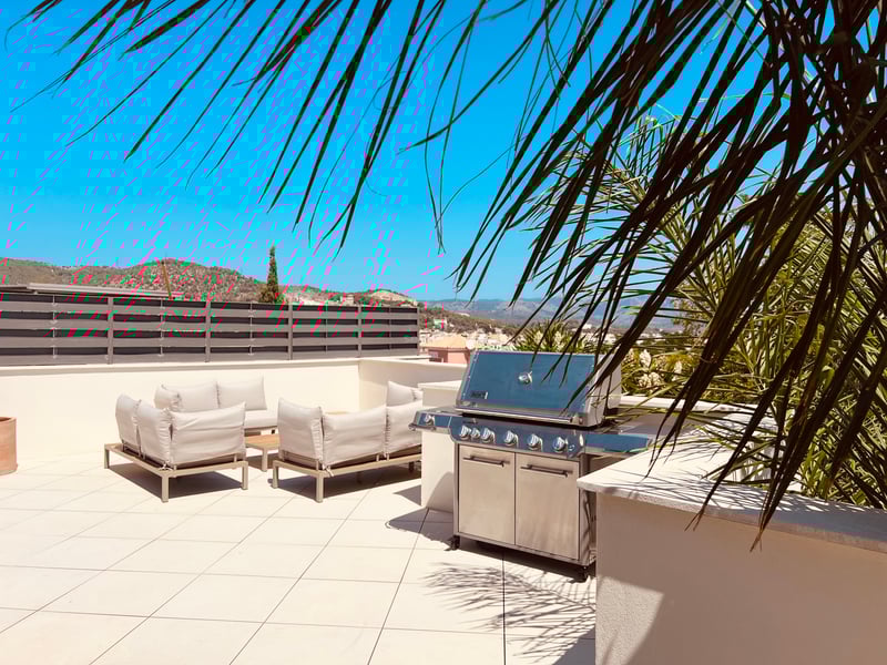 Toit-terrasse ensoleillé avec grand barbecue en inox, salon de jardin moderne et vue sur les montagnes.