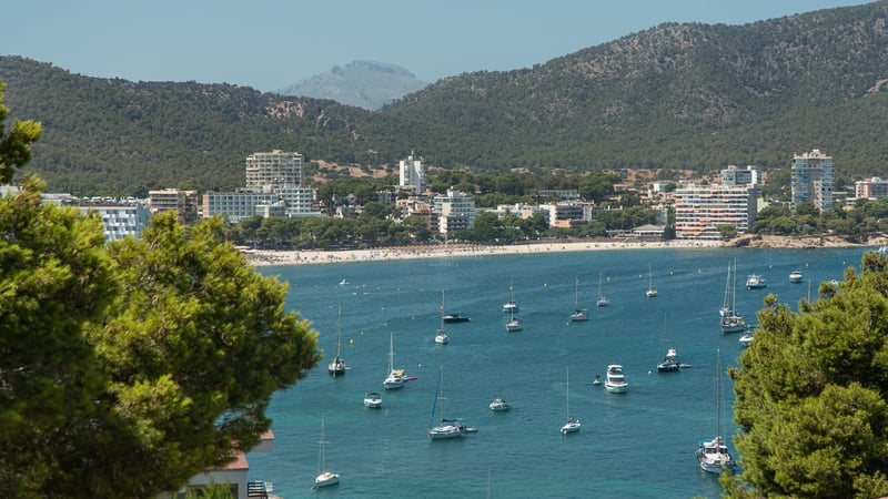 Vue surélevée d'une baie turquoise avec des voiliers, une plage de sable et des montagnes vertes sous un ciel dégagé.