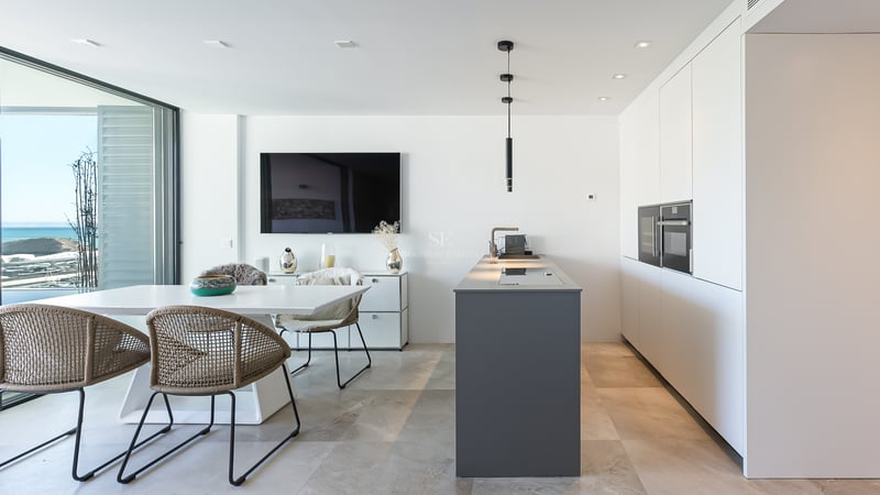 Moderne weiße Küche mit grauer Insel, Esstisch und bodentiefen Fenstern mit Blick auf das Mittelmeer.
