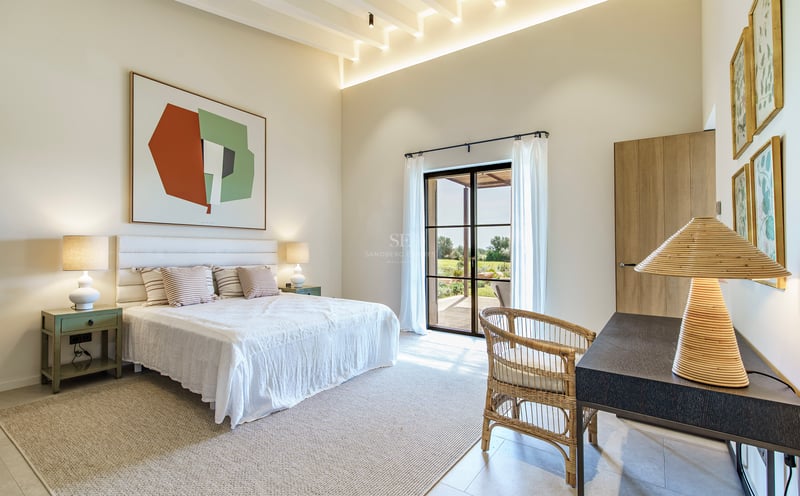 Helles, modernes Schlafzimmer mit hohen Decken, weißem Bett, Korbstuhl und großen Glastüren zum Garten.