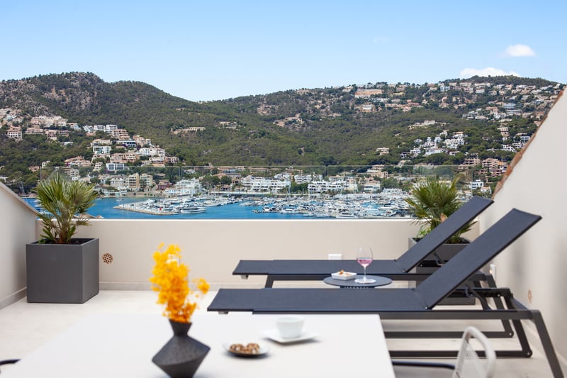 Sun loungers on a modern terrace overlooking a harbor with white boats and lush green hills under a clear blue sky.
