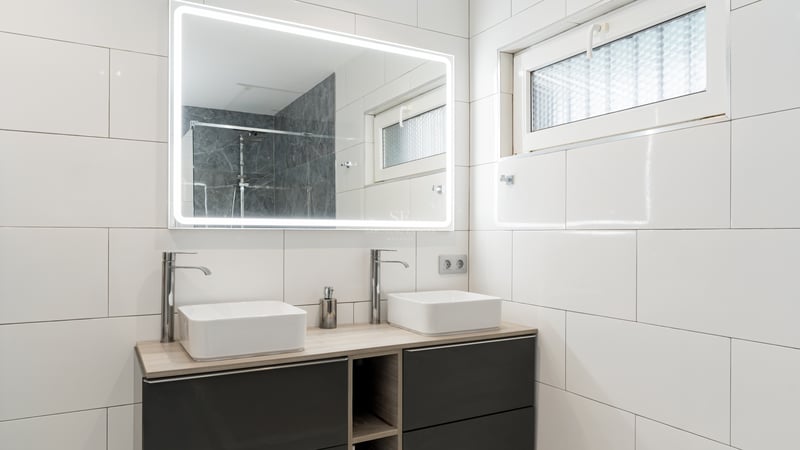 Salle de bain moderne avec deux vasques carrées, un grand miroir éclairé et des murs en carrelage blanc.