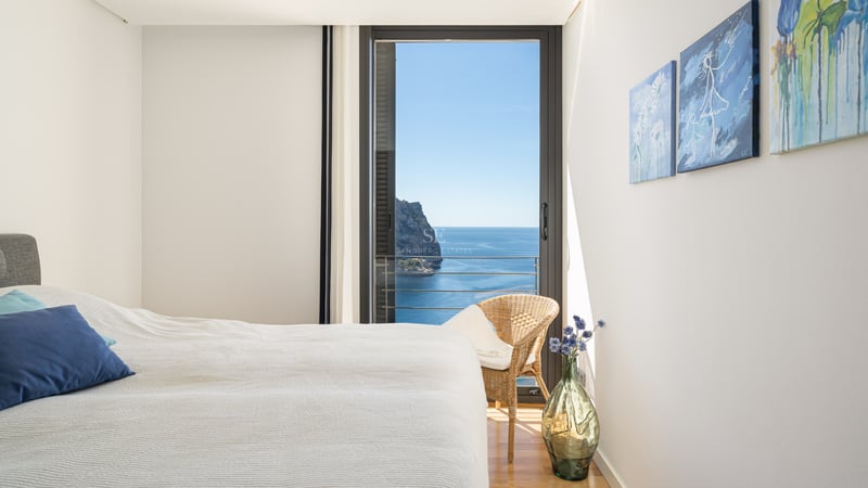 Bright bedroom with white linens, blue accent pillow, and floor-to-ceiling glass doors overlooking the Mediterranean sea.