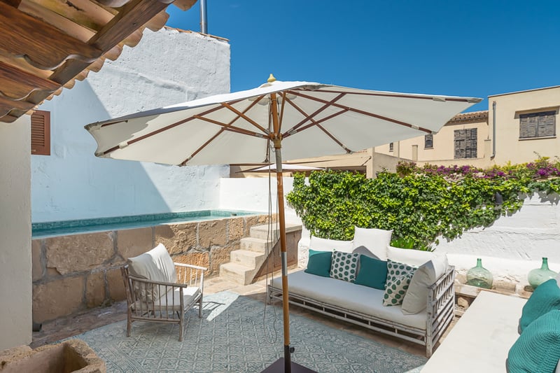 Patio méditerranéen ensoleillé avec piscine en pierre surélevée, canapé blanc et parasol.