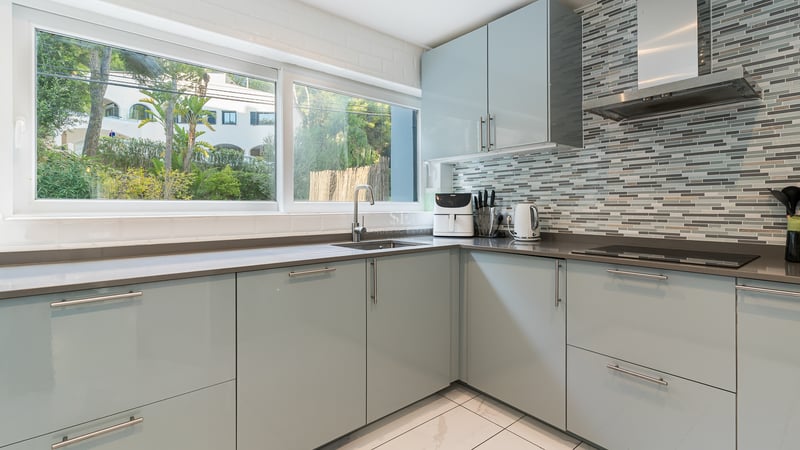 Cuisine moderne avec meubles bleu clair, plans de travail gris, grande fenêtre et crédence en mosaïque.