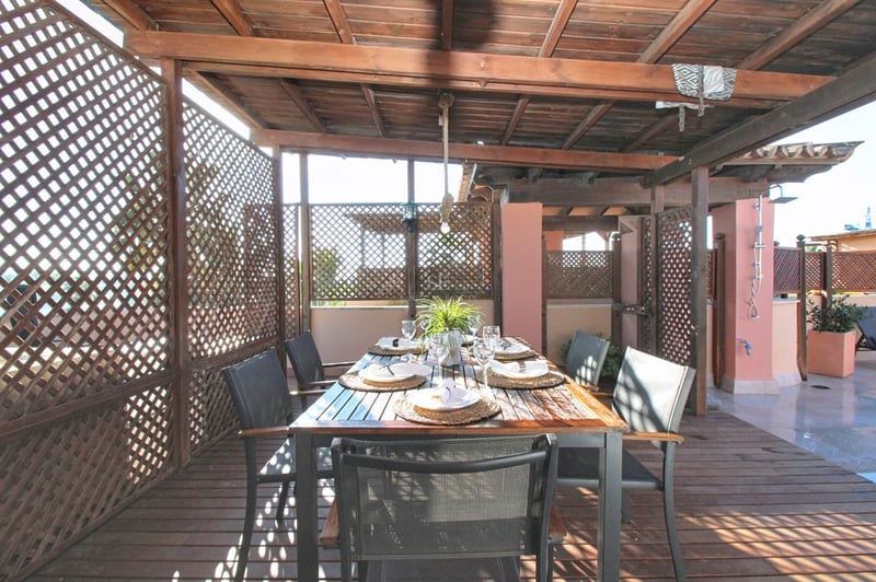 Matsalsbord under en träpergola med insynsskydd och trädäck på en solig terrass.