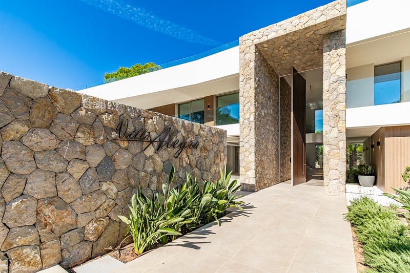 Entrée de villa moderne avec mur en pierre naturelle, enseigne Villa Alegra et grande porte pivotante en bois.