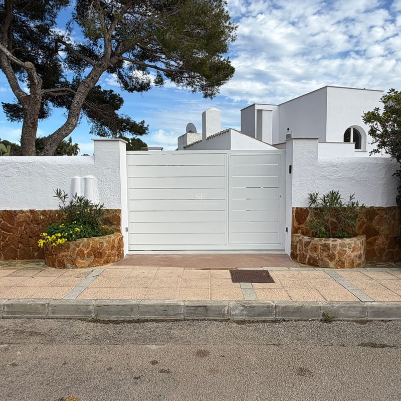 Large white metal gate flanked by white stucco and brown stone walls under a blue sky with a pine tree.