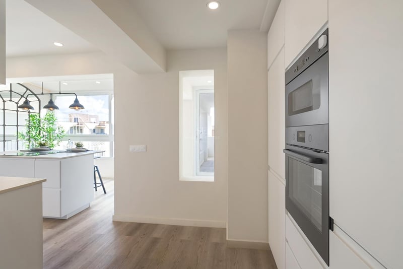 Moderne weiße Küche mit schwarzen Einbaubacköfen, Holzboden und weißer Insel unter Industrie-Leuchten.