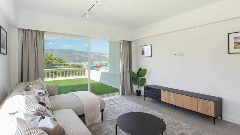 Salon moderne avec canapé gris et baies vitrées ouvrant sur une terrasse vue mer et montagne.