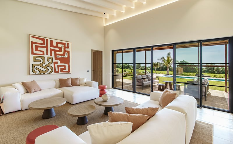 Heller Wohnraum mit weißen Sofas, Holztischen und bodentiefen Fenstern mit Blick auf Pool und Garten.