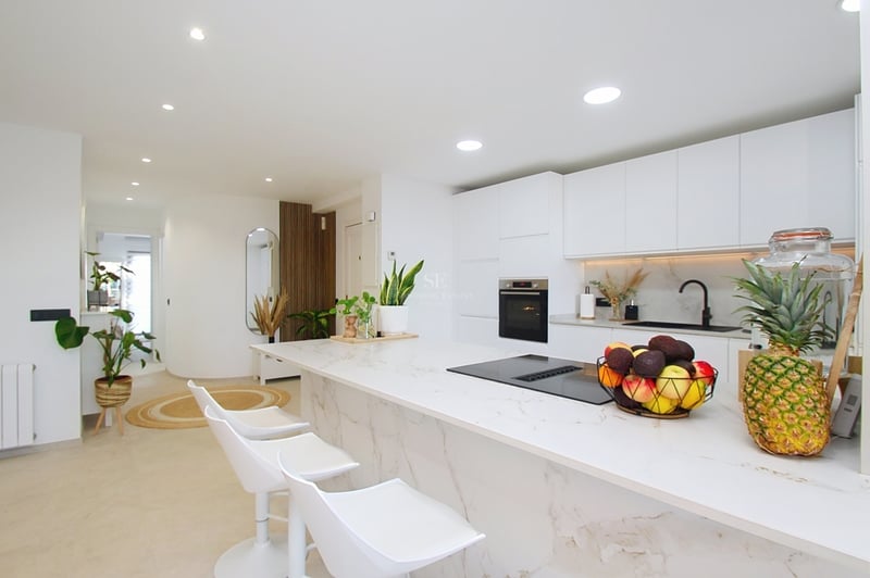 Minimalistiskt vitt kök med marmorliknande köksö, vita barstolar och inbyggda vitvaror.