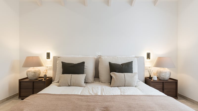 Lit avec tête de lit rembourrée, coussins décoratifs, tables de chevet en bois et lampes design à lumière chaude.