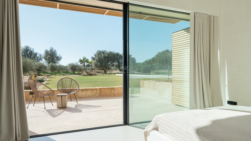 Utsikt från ett ljust sovrum genom skjutdörrar i glas mot en solig terrass och grönskande trädgård.