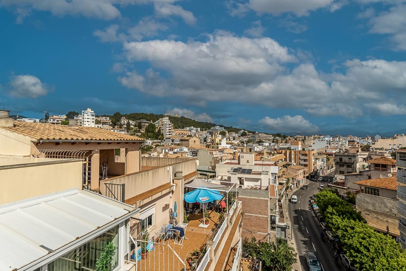 Erhöhte Ansicht mediterraner Hausdächer, einer von Bäumen gesäumten Straße und ferner Berge unter blauem Himmel.